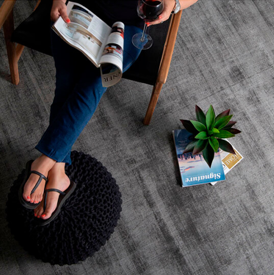 Lady sitting on a black and wooden armchair with feet up on a black pouf enjoying a glass of red wine and reading a magazine